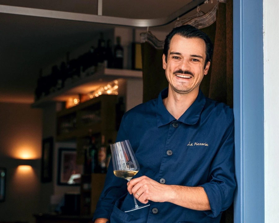 <think> Got it, let's create the alt text. First, identify what's visible. A man in a blue shirt with "La Pizzeria" on it ho…