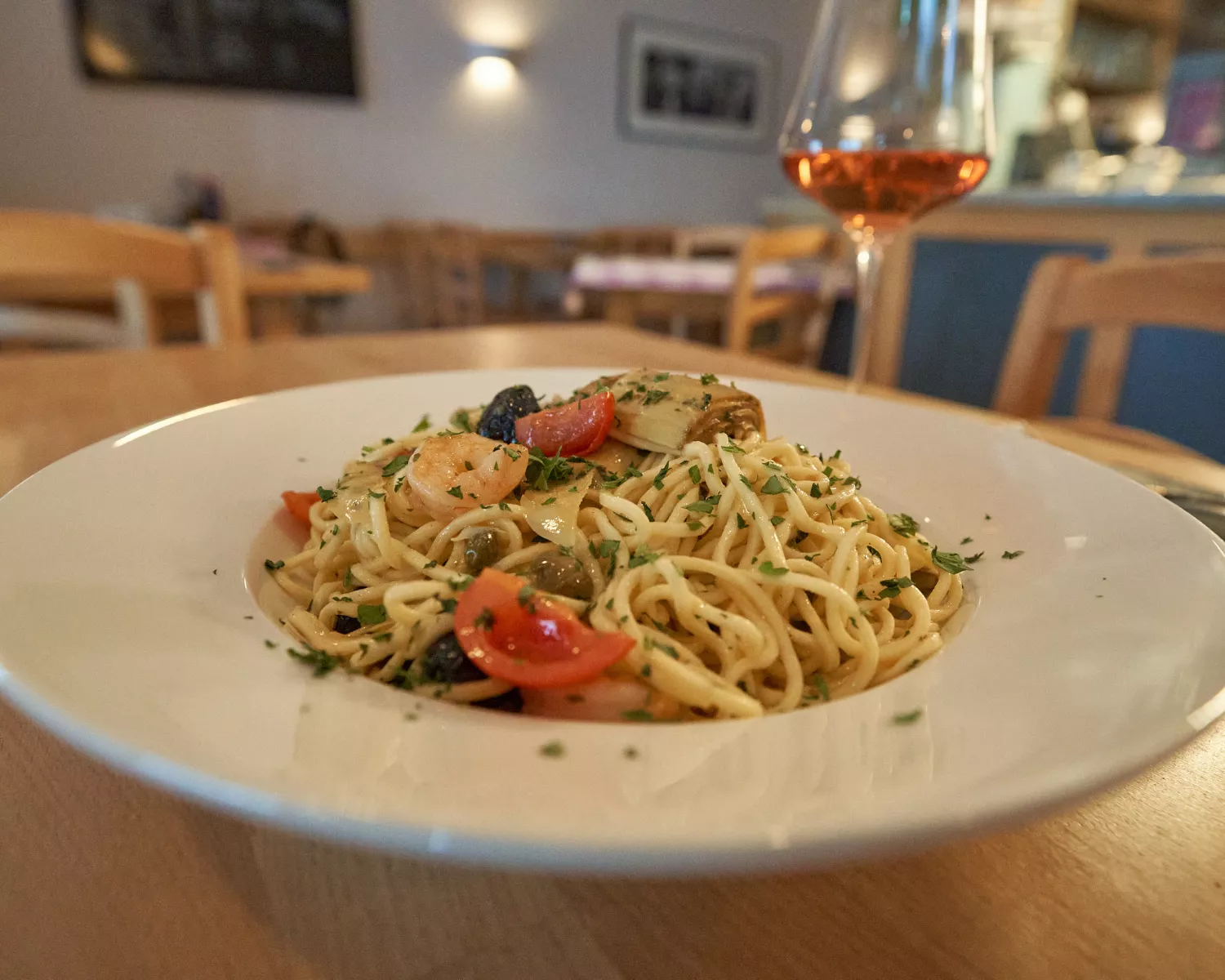 <think> Got it, let's create the alt text. The image shows a plate of pasta with shrimp, tomatoes, olives, and herbs on a wo…