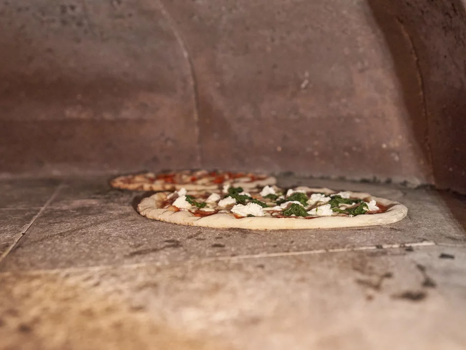 <think> Got it, let's write the alt text. The image shows two pizzas inside a stone oven. The main pizza in focus has toppin…