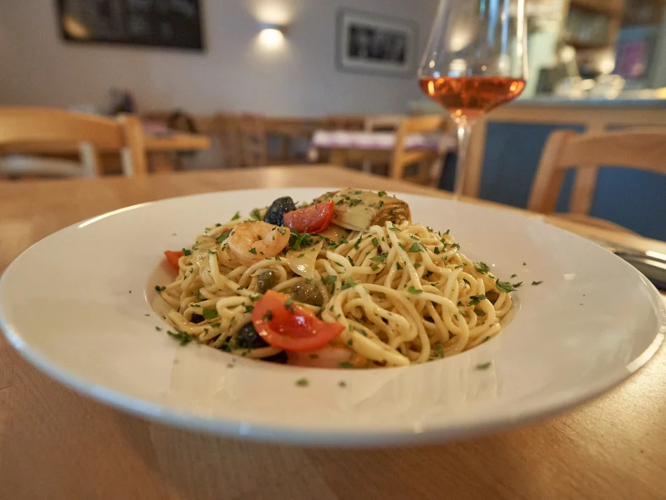 <think> Got it, let's create the alt text. The image shows a plate of pasta with shrimp, tomatoes, olives, and herbs on a wo…