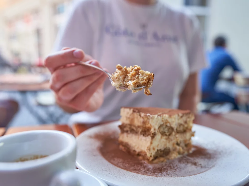 <think> Got it, let's create the alt text. First, identify what's visible. There's a person holding a spoon with tiramisu, a…